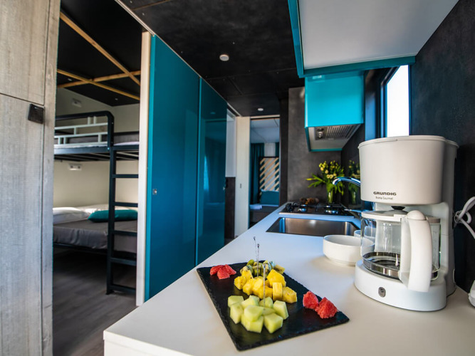 Moderne chaletkeuken met koffiemachine, schotel met vers fruit en stapelbedden op de achtergrond.