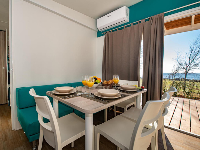 Sala da pranzo in chalet moderno apparecchiata per quattro, bevande fresche e vista mare dalla finestra.