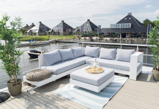 Terras met moderne loungeset aan het water, uitzicht op huisjes, boten en een vakantiepark in de zomer.