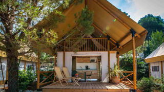 Twee slaapkamer glamping safari loft tent met houten veranda, stoelen en omgeven door groene natuur.