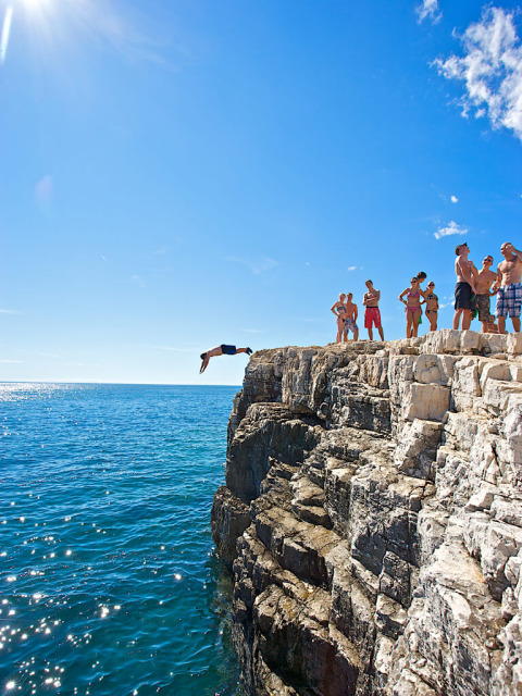Group of people at Camping Arena Stoja in Istria, Croatia, watching someone cliff jump into the sparkling sea.