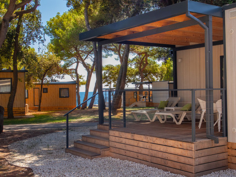 Deluxe Camping Villa Sea View, terrazza moderna con lettini, circondata da alberi e vista parziale sul mare.
