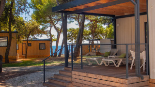Deluxe Camping Villa Sea View, moderne veranda met ligstoelen, omgeven door bomen en zicht op zee.