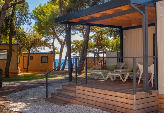 Deluxe Camping Villa Sea View, moderne veranda met ligstoelen, omgeven door bomen en zicht op zee.
