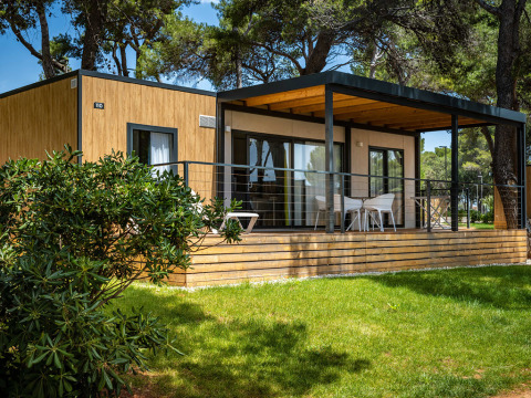 Moderne Lodge bei Camping Villa mit Holzfassade, großer Veranda, Sitzbereich und viel Grün.