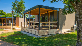 Modern lodge with spacious wooden deck, surrounded by green lawn and trees on a sunny day.