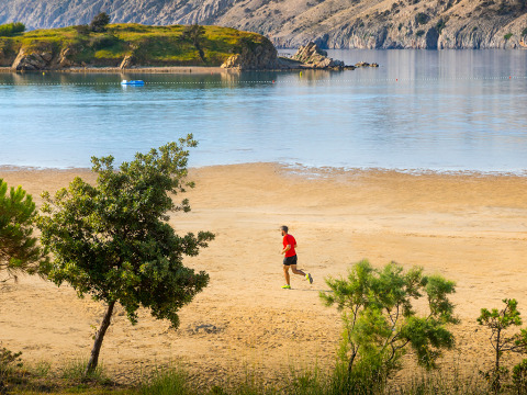 Uomo che corre sulla spiaggia sabbiosa con alberi verdi e mare calmo a Valamar Camping San Marino, Croazia.
