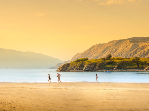 Spiaggia vicino a Lopar, Croazia, con tre persone sulla sabbia, acqua calma e montagne illuminate.