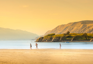 Spiaggia vicino a Lopar, Croazia, con tre persone sulla sabbia, acqua calma e montagne illuminate.