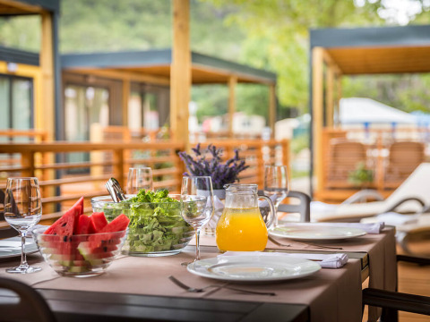 Outdoor table at a lodge set with watermelon, salad, juice, and glasses, ready for a summer meal.