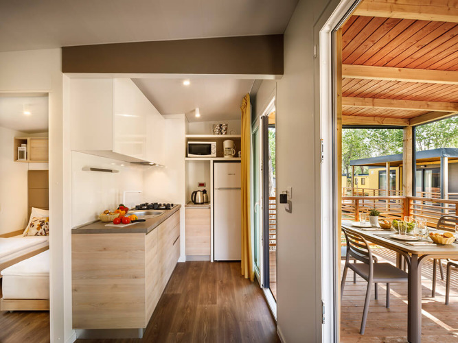Cuisine moderne de lodge avec porte ouverte sur terrasse couverte, table extérieure et vue sur d'autres chalets.