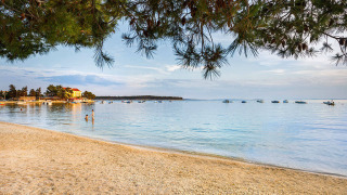Peaceful beach with calm sea, boats, and pine branches near Banjol, Primorje-Gorski Kotar, Croatia.