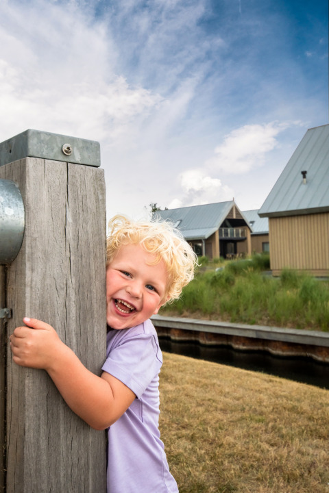 Een jong kind met blond krullend haar lacht en omhelst een houten paal bij Oesterdam Resort in Zeeland.
