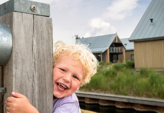 Een jong kind met blond krullend haar lacht en omhelst een houten paal bij Oesterdam Resort in Zeeland.