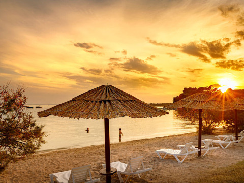 Playa al atardecer cerca de Banjol, Croacia, con sombrillas de paja, tumbonas y un cielo dorado.