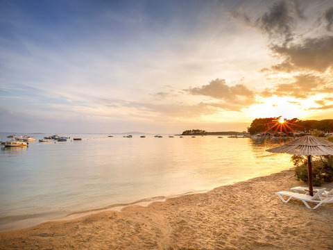 Coucher de soleil sur la plage de Valamar Camping Padova avec transats, parasol et bateaux sur l'eau calme de Croatie.