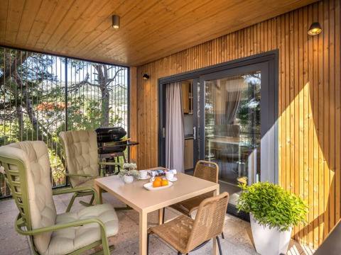 Covered patio at Hilltop Green Chalet with cushioned chairs, dining table, plants, and a grill.