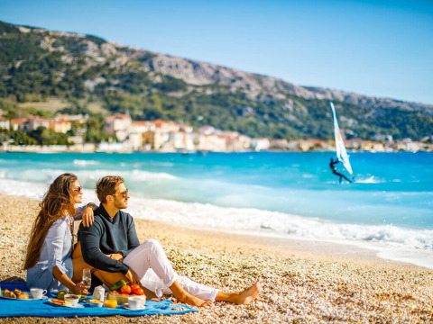 Par nyder en picnic på stranden i nærheden af Baška i Kroatien med udsigt til hav og windsurfer.