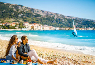 Pareja disfruta de un picnic en la playa cerca de Baška, Croacia, con vista al mar y a un windsurfista.