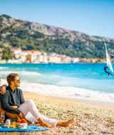 Paar genießt Picknick am Strand nahe Baška, Kroatien, mit Blick auf das Meer und einen Windsurfer.