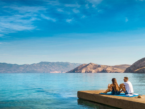 Couple relaxing on a jetty overlooking clear blue water and mountains at Valamar Camping Baška, Croatia.