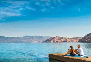 Couple relaxing on a jetty overlooking clear blue water and mountains at Valamar Camping Baška, Croatia.