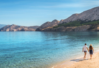 Coppia che cammina su una spiaggia tranquilla con acqua limpida e montagne sullo sfondo in una giornata di sole.
