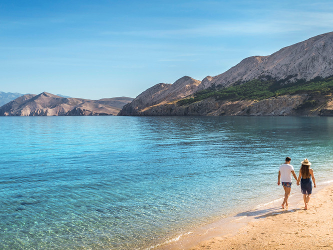 Couple marchant sur une plage paisible avec de l’eau claire et des montagnes en arrière-plan sous le soleil.
