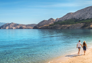 Pareja caminando por una playa tranquila con agua cristalina y montañas al fondo bajo un cielo despejado.