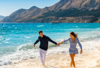 Ein Paar spaziert lachend am Strand von Valamar Camping Baška, Kroatien, mit Bergen im Hintergrund.