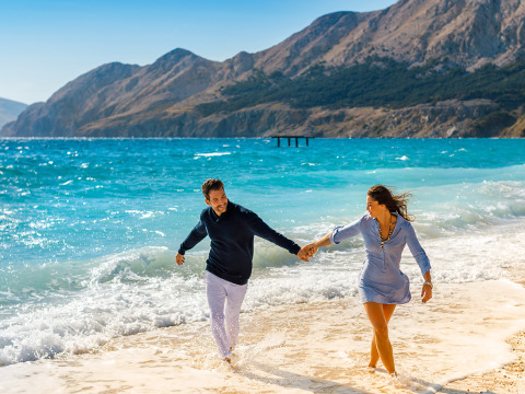 A couple walks hand in hand along the beach at Valamar Camping Baška, Croatia, with mountains behind them.