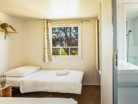 Lichte slaapkamer met twee eenpersoonsbedden, raam, badkamer en kleurrijk gordijn bij Camping Brioni, Kroatië.