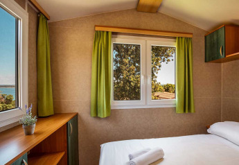 Slaapkamer in chalet Brioni Home Seaside bij Camping Brioni in Kroatië met groene gordijnen en buitenzicht.