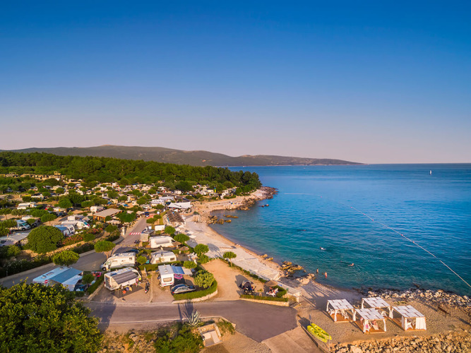 Vista aérea de un lodge costero con caravanas y tiendas junto al mar, rodeado de bosque y agua cristalina.