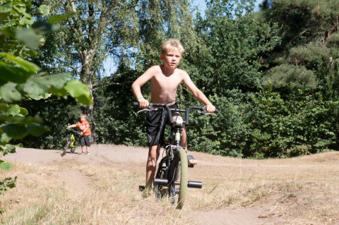 Twee kinderen fietsen op een zandpad omgeven door bomen bij een vakantiepark met glampingaccommodaties.
