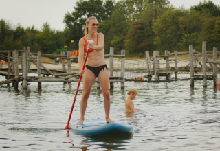 Vrouw peddelt op een supboard in het water bij een vakantiepark met kinderen en houten brug op de achtergrond.