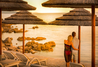 Coppia che guarda il tramonto sotto gli ombrelloni sulla spiaggia vicino a Krk, Primorje-Gorski Kotar, Croazia.