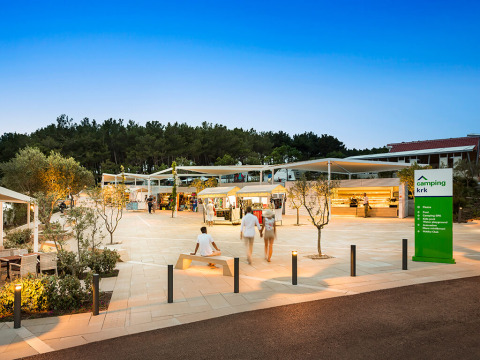 Soirée au camping Valamar Krk en Croatie, avec des visiteurs, stands et arbres autour de la place.