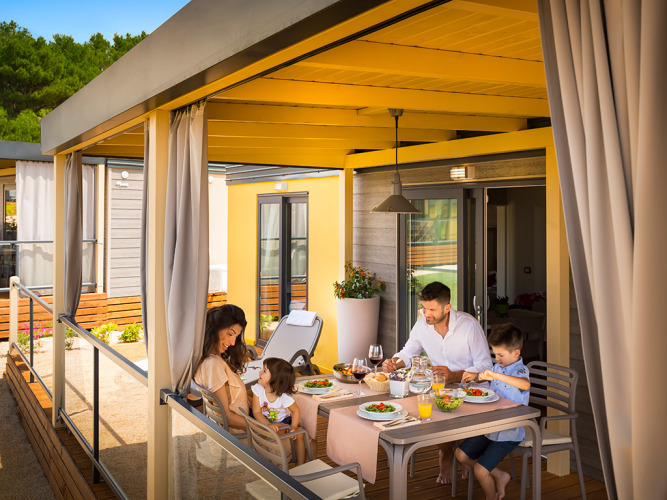 Familie nyder måltid på overdækket terrasse ved Bella Vista Home Lodge omgivet af natur.