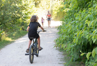 Kind fietst op een zandpad langs groene struiken in een vakantiepark met glamping, familie wandelt verderop