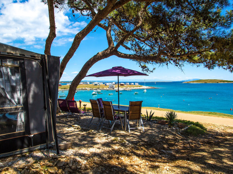 Camping Arena Stupice in Istria, Croazia con tavolo all’ombra, vista mare blu e barche ancorate vicino.