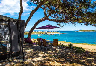 Camping Arena Stupice in Istria, Croazia con tavolo all’ombra, vista mare blu e barche ancorate vicino.