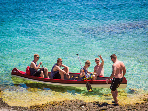 Five men relaxing in a canoe on clear blue water near Premantura, Istria, Croatia, by the rocky shore.