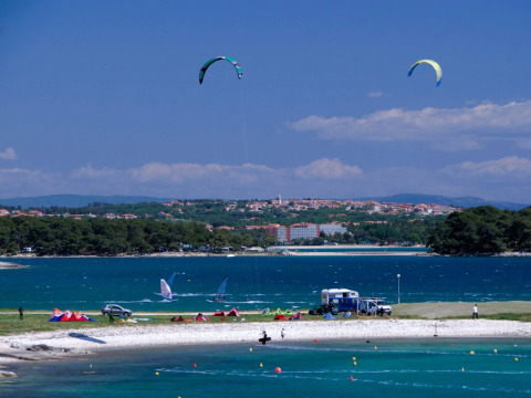 Wassersport und Camping am Strand von Arena Stupice in Istrien, Kroatien, mit Stadt im Hintergrund.