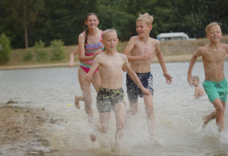 Kinderen en een vrouw rennen lachend door het water bij een vakantiepark met glamping aan een meer.