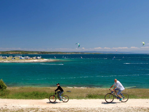 Due persone pedalano lungo la costa al Camping Arena Stupice, con mare, windsurfisti e camper sullo sfondo.