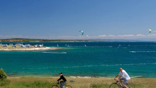 To personer cykler langs kysten ved Camping Arena Stupice, med hav, windsurfere og campingvogne i baggrunden.