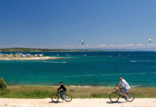 Twee mensen fietsen aan de kust bij Camping Arena Stupice, met zee, windsurfers en campers op de achtergrond.