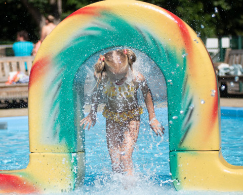 Een jong meisje speelt in een waterpark bij een glamping accommodatie, lachend onder een sproeiende boog.
