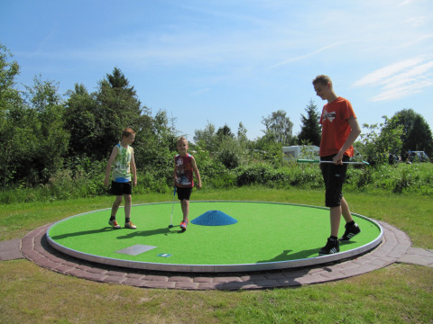 Kinderen spelen minigolf op een glamping vakantiedomein, met groene natuur en blauwe lucht op de achtergrond.